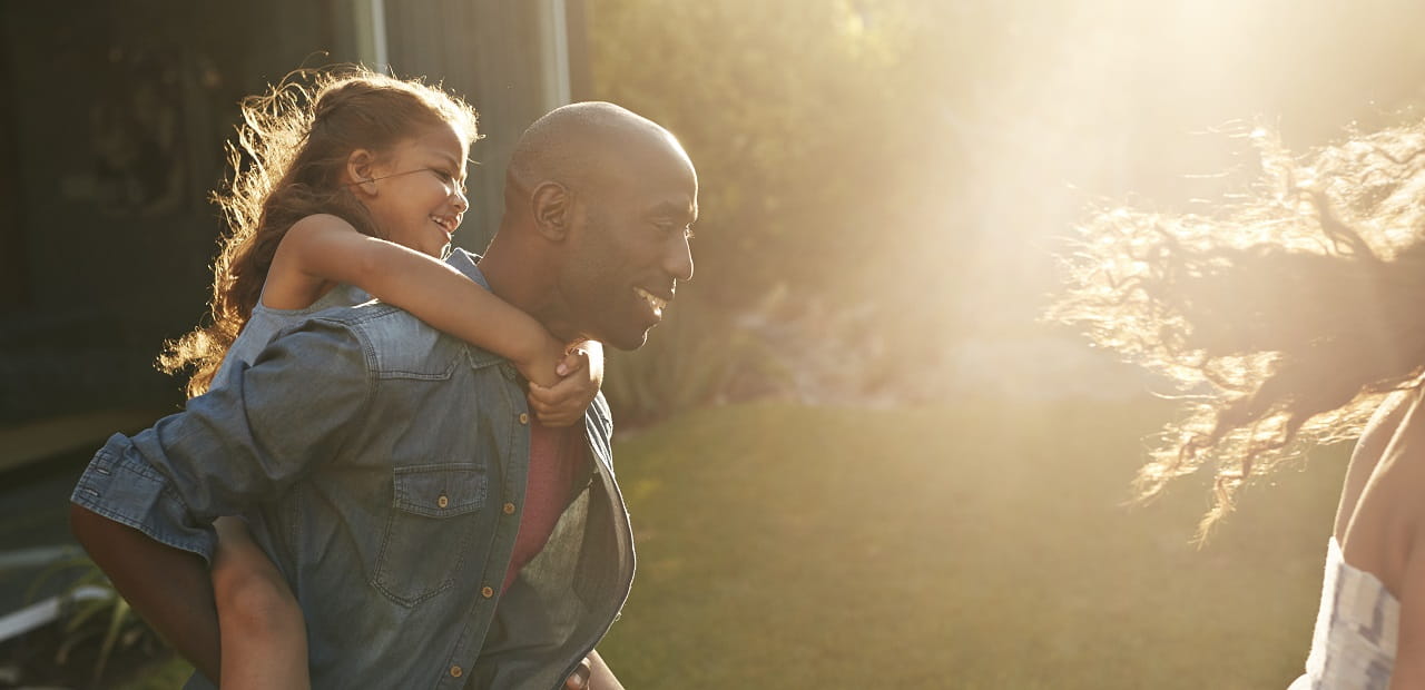 Man plays with his family in a backyard with his daughter on his back. 