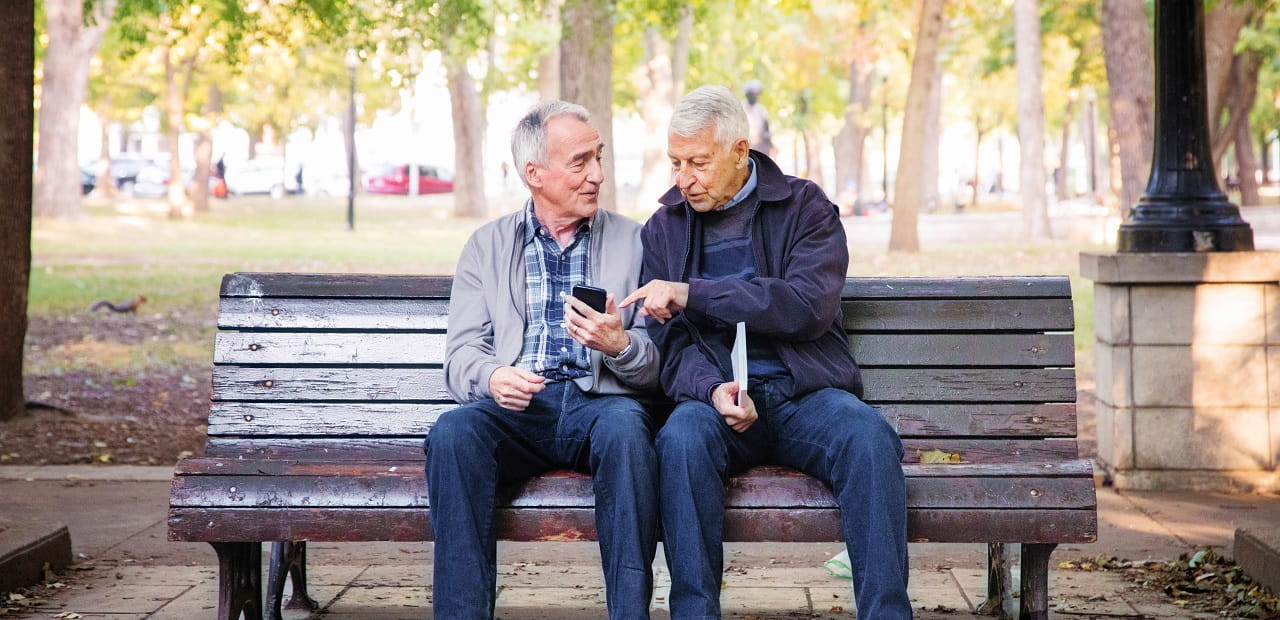 An elderly same sex couple sits on a park bench talking.