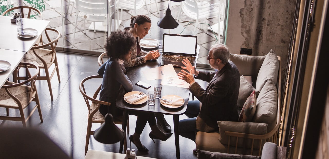 An overhead shot of three professionals sitting at a café table together. A few papers and an open laptop are visible on the table.