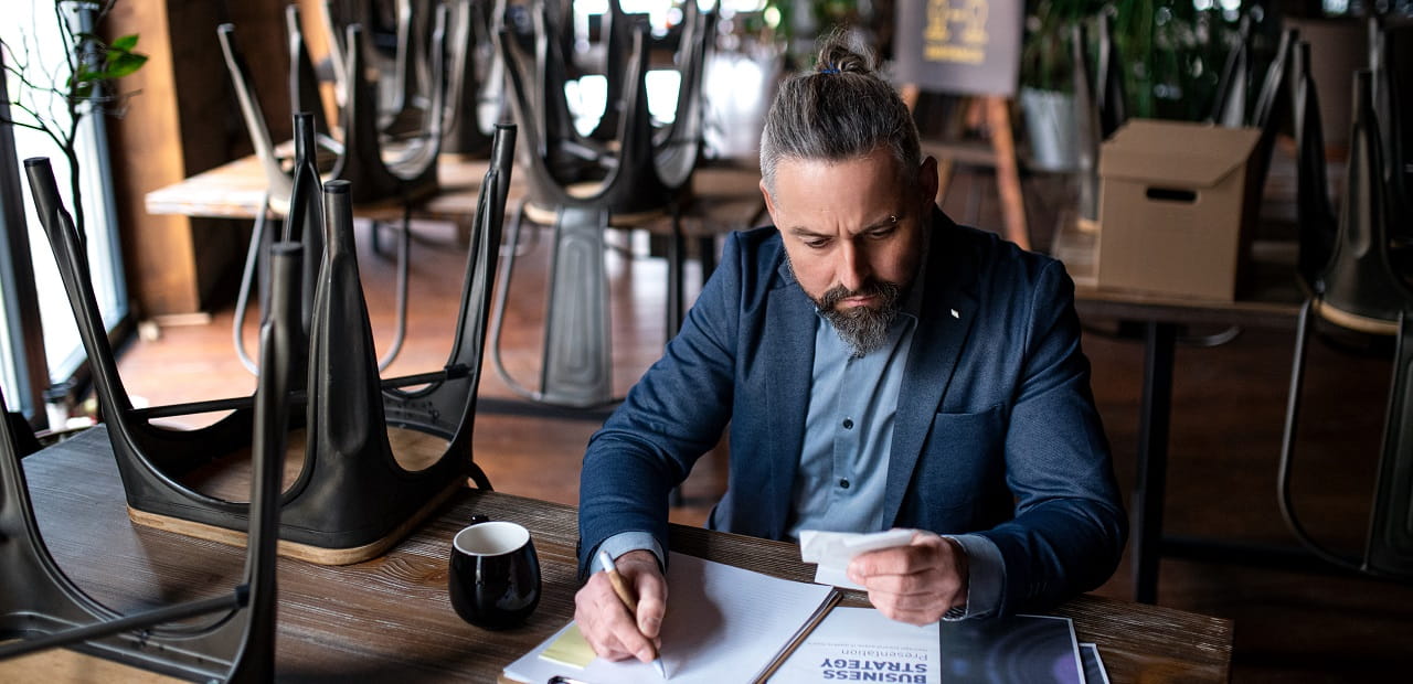 Man sits at a table in a closed restaurant writing on a sheet of paper and looking at a receipt.